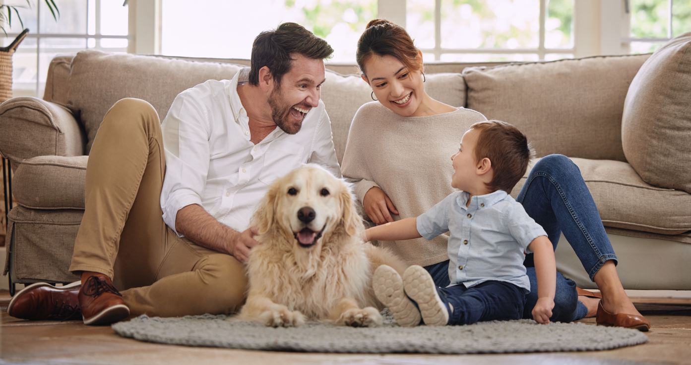 A happy family of three (mother, father, and young son) sitting on the living room floor with their smiling golden retriever.